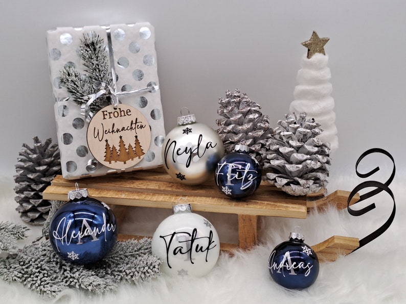 Pode incluir: Um conjunto de seis enfeites de Natal personalizados com detalhes prateados e azuis. Os enfeites s&atilde;o decorados com nomes e flocos de neve. Os enfeites s&atilde;o exibidos em um tren&oacute; de madeira com uma base de pele sint&eacute;tica branca. Os enfeites s&atilde;o cercados por pinhas e uma pequena &aacute;rvore de Natal artificial branca.