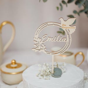 Pode incluir: Um bolo branco de dois andares com um topo de bolo de madeira. O topo &eacute; um c&iacute;rculo com o nome "Emilia" e uma pomba com o texto "Zur Konfirmation".