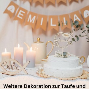 Pode incluir: Um bolo branco de dois andares com um topo de bolo de madeira que diz "Taufe Emilia" em letras douradas. O bolo &eacute; decorado com flores brancas e vegeta&ccedil;&atilde;o. O bolo est&aacute; sobre um suporte de bolo de madeira. H&aacute; velas e um peixe de madeira com o nome "Emilia" no fundo. O texto "Weitere Dekoration zur Taufe und Kommunion im Shop erh&auml;ltlich" est&aacute; na parte inferior da imagem. O endere&ccedil;o do site "WWW.ROXISKREATIVSHOP.DE" est&aacute; abaixo do texto.