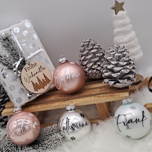 Pode incluir: Quatro enfeites de Natal personalizados com os nomes "Edda", "Katharina", "Frank" e "Silvia" escritos em letras brancas. Os enfeites est&atilde;o em um tren&oacute; de madeira com um presente embrulhado em papel branco com bolinhas prateadas e uma etiqueta de madeira que diz "Frohe Weihnachten" com um desenho de &aacute;rvore de Natal.