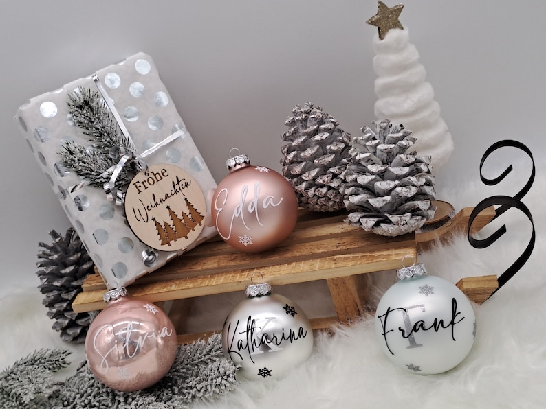 Pode incluir: Quatro enfeites de Natal personalizados com os nomes "Edda", "Katharina", "Frank" e "Silvia" escritos em letras brancas. Os enfeites est&atilde;o em um tren&oacute; de madeira com um presente embrulhado em papel branco com bolinhas prateadas e uma etiqueta de madeira que diz "Frohe Weihnachten" com um desenho de &aacute;rvore de Natal.