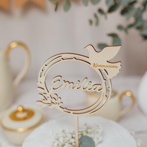 Pode incluir: Um topo de bolo de madeira com uma pomba e uma coroa, o nome "Emilia" e a palavra "Comunh&atilde;o" est&atilde;o inscritos no topo.
