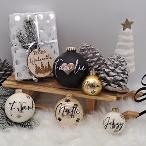 Pode incluir: Um conjunto de seis enfeites de Natal com nomes e designs personalizados. Os enfeites s&atilde;o brancos, pretos, dourados e creme com letras douradas. Os enfeites s&atilde;o decorados com flocos de neve e um cora&ccedil;&atilde;o. Os enfeites est&atilde;o dispostos em um tren&oacute; de madeira com uma caixa de presente branca e uma &aacute;rvore de Natal branca no fundo.