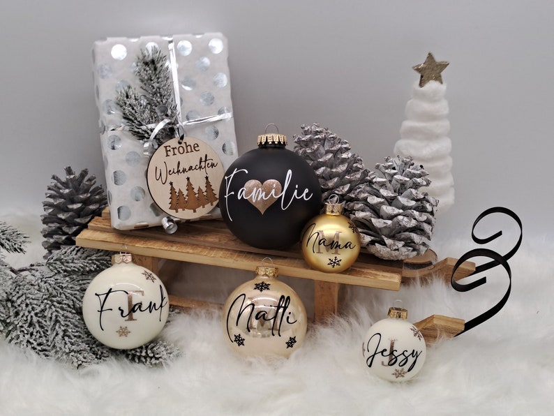 Pode incluir: Um conjunto de seis enfeites de Natal com nomes e designs personalizados. Os enfeites s&atilde;o brancos, pretos, dourados e creme com letras douradas. Os enfeites s&atilde;o decorados com flocos de neve e um cora&ccedil;&atilde;o. Os enfeites est&atilde;o dispostos em um tren&oacute; de madeira com uma caixa de presente branca e uma &aacute;rvore de Natal branca no fundo.