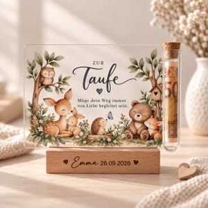 Könnte beinhalten: Eine rechteckige Acrylplatte auf einem Holzsockel mit Waldtierillustrationen von Eule, Reh, Hase, Igel, Bär und Fuchs. Der Text lautet "ZUR Taufe" und "Möge dein Weg immer von Liebe begleitet sein." Der Sockel ist mit "Emma 26.09.2026" graviert und enthält ein Glasreagenzglas mit Korken, in dem sich ein 50-Euro-Schein befindet.