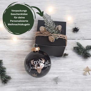 Pode incluir: Enfeite de Natal preto com glitter dourado e o texto "Frohe Weihnachten" em branco. O enfeite est&aacute; embalado em uma caixa preta com um la&ccedil;o de corda marrom e um pequeno ramo de vegeta&ccedil;&atilde;o.