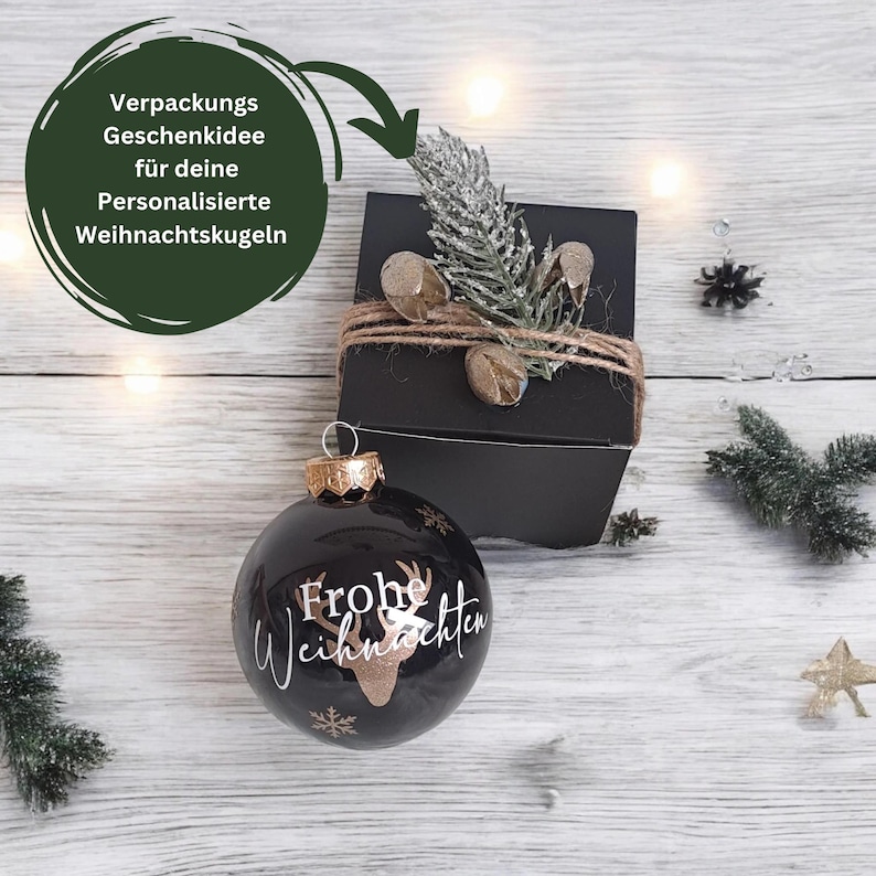 Pode incluir: Enfeite de Natal preto com glitter dourado e o texto "Frohe Weihnachten" em branco. O enfeite est&aacute; embalado em uma caixa preta com um la&ccedil;o de corda marrom e um pequeno ramo de vegeta&ccedil;&atilde;o.