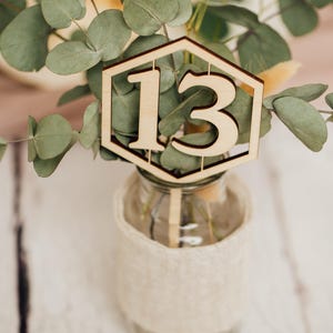 May include: A wooden table number with the number 13 cut out in the center. The number is placed in a small glass vase with a burlap wrap and greenery.