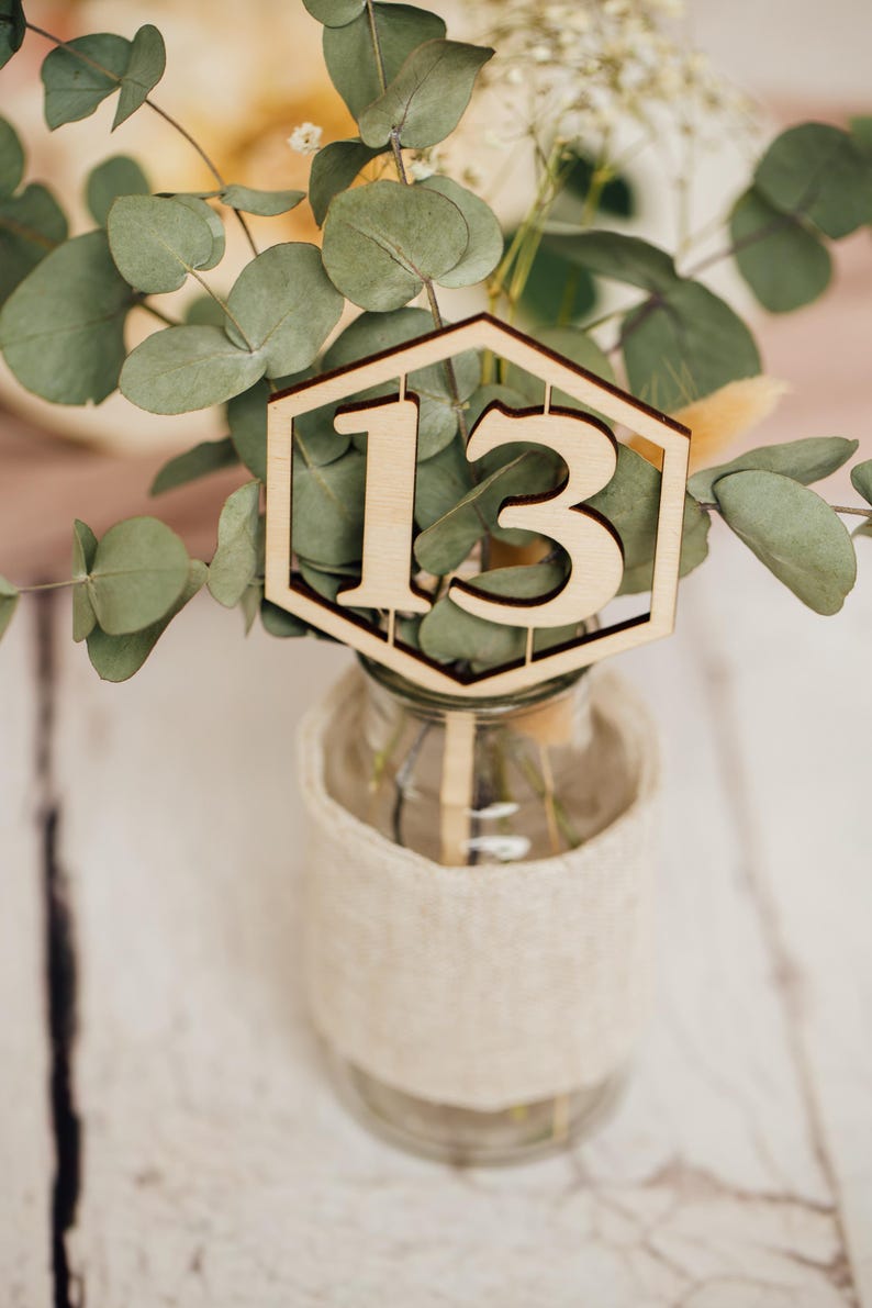 May include: A wooden table number with the number 13 cut out in the center. The number is placed in a small glass vase with a burlap wrap and greenery.