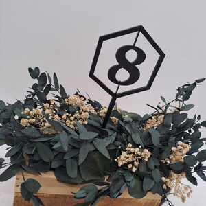 May include: A black hexagonal table number with the number 8 inside. The table number is placed in a wooden box filled with green eucalyptus leaves and small white flowers.