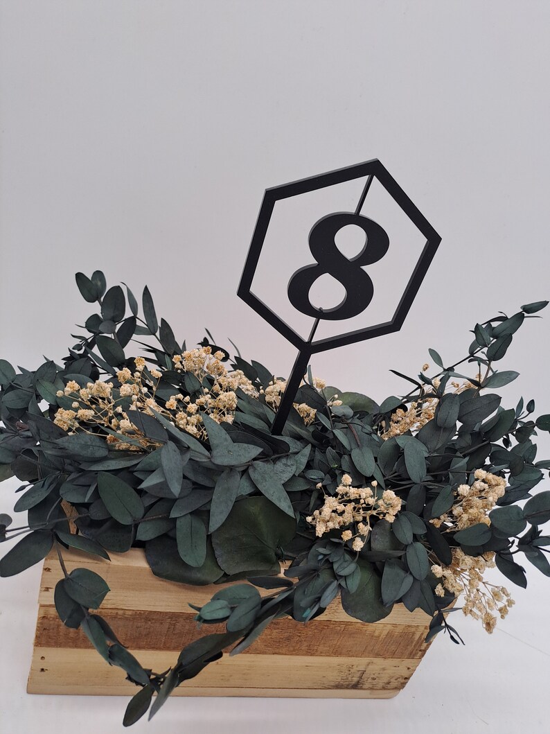 May include: A black hexagonal table number with the number 8 inside. The table number is placed in a wooden box filled with green eucalyptus leaves and small white flowers.