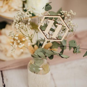 May include: A wooden table number in the shape of a hexagon with the number "11" cut out. The table number is placed in a small glass vase with white baby's breath and eucalyptus.