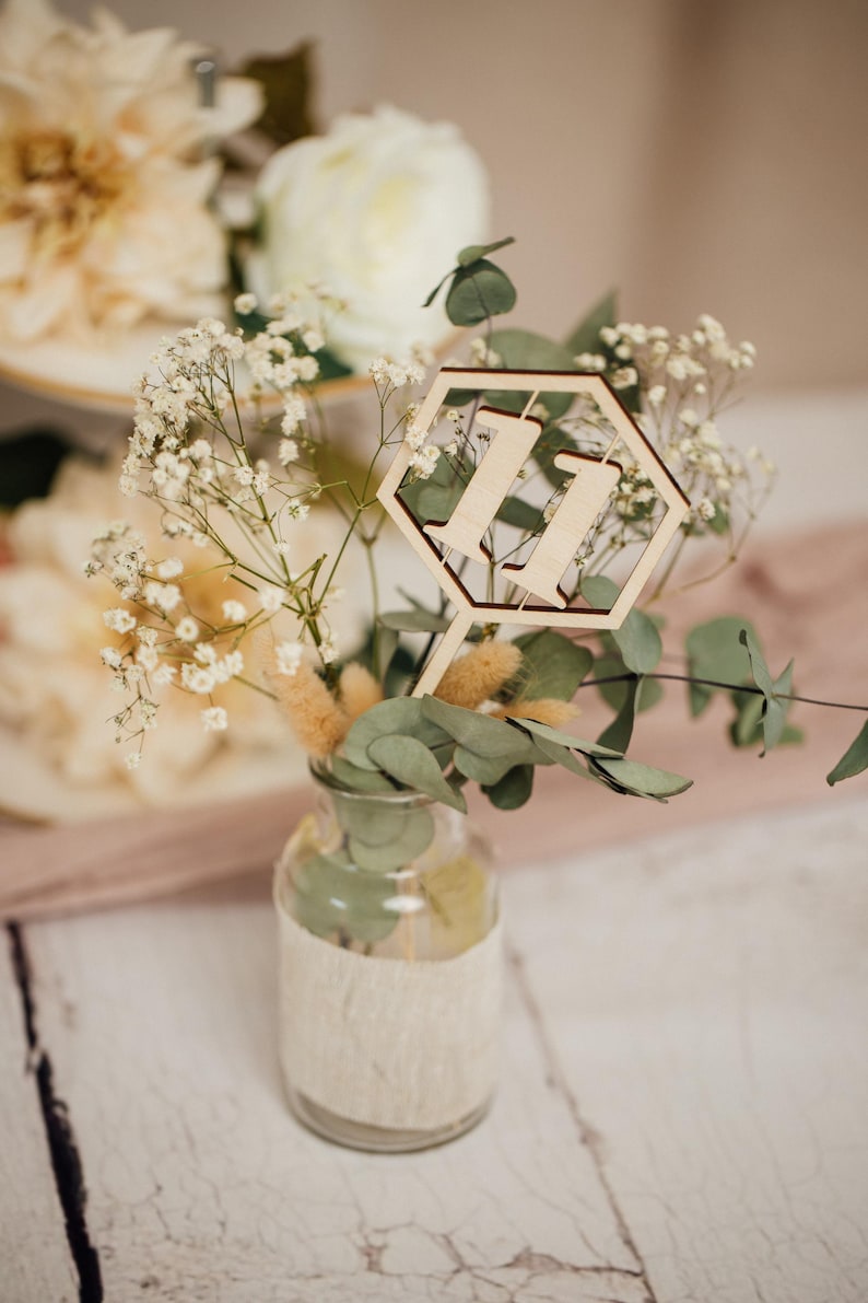 May include: A wooden table number in the shape of a hexagon with the number "11" cut out. The table number is placed in a small glass vase with white baby's breath and eucalyptus.