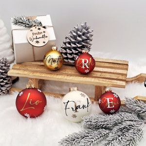 Pode incluir: Cinco enfeites de Natal personalizados em um tren&oacute; de madeira. Os enfeites s&atilde;o vermelhos, dourados e brancos com letras douradas. Os nomes nos enfeites s&atilde;o Matt, Lio, Frank e Emi. O tren&oacute; est&aacute; sobre uma superf&iacute;cie branca com uma pequena &aacute;rvore de pinho e pinhas.