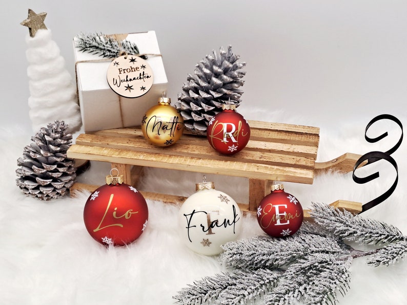Pode incluir: Cinco enfeites de Natal personalizados em um tren&oacute; de madeira. Os enfeites s&atilde;o vermelhos, dourados e brancos com letras douradas. Os nomes nos enfeites s&atilde;o Matt, Lio, Frank e Emi. O tren&oacute; est&aacute; sobre uma superf&iacute;cie branca com uma pequena &aacute;rvore de pinho e pinhas.
