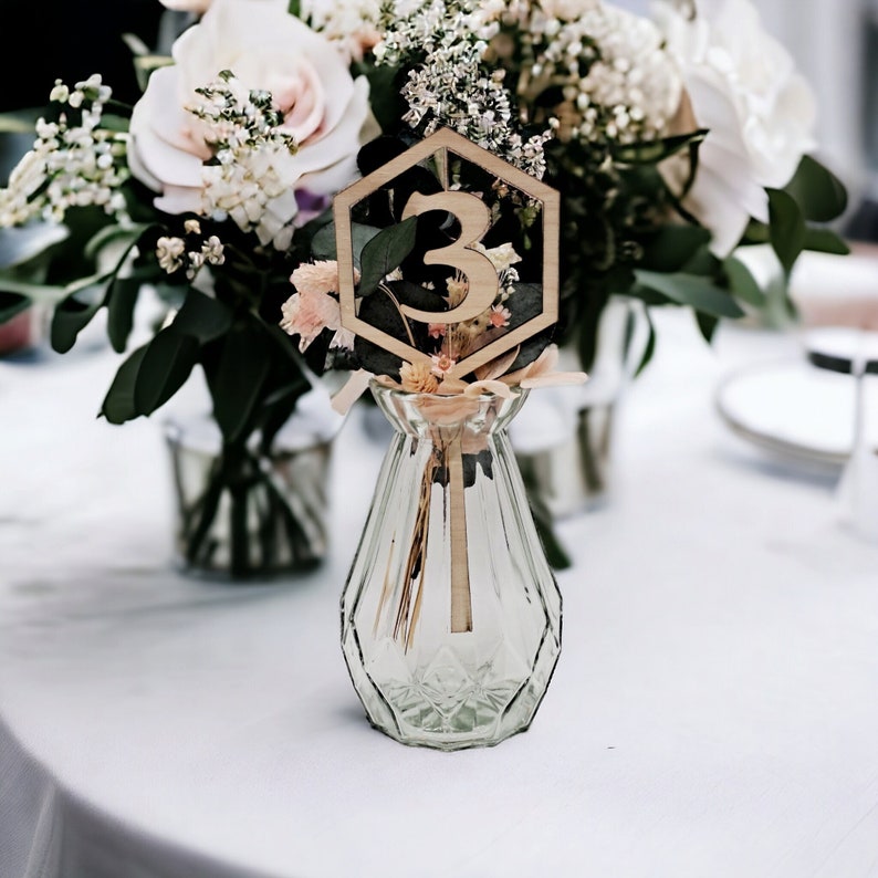 May include: A wooden table number with a geometric hexagon shape and the number "3" in the center. The table number is placed in a clear glass vase with dried flowers.