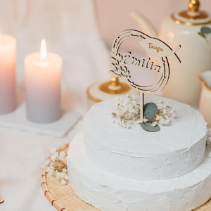 Pode incluir: Um bolo branco de dois andares com cobertura branca &eacute; decorado com um topo de bolo de madeira que diz "Taufe Emilia". O bolo est&aacute; em um suporte de bolo de bambu tecido.