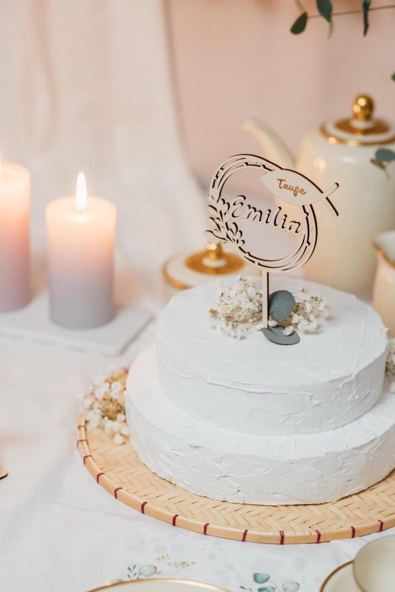 Pode incluir: Um bolo branco de dois andares com cobertura branca &eacute; decorado com um topo de bolo de madeira que diz "Taufe Emilia". O bolo est&aacute; em um suporte de bolo de bambu tecido.