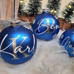 Pode incluir: Tr&ecirc;s enfeites de Natal azuis com flocos de neve brancos e letras prateadas. Os enfeites s&atilde;o personalizados com os nomes "Ben", "Karl" e "Maxim".