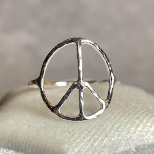 May include: A silver-colored ring featuring the peace symbol. The ring's design is a classic peace sign, with a circular shape and three lines. The ring is set against a soft, light-colored background.