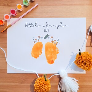May include: A white card with the text "Ottavie's pumpkin 2023" and two orange handprints in the shape of a pumpkin. A white string with yellow and white pom-poms hangs below the card.