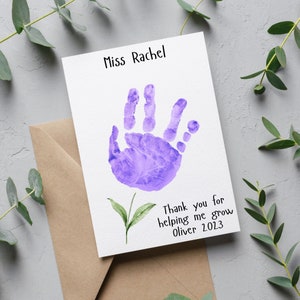 May include: A white card with a purple handprint and a green leaf stem. The card says "Miss Rachel" at the top and "Thank you for helping me grow Oliver 2023" at the bottom.
