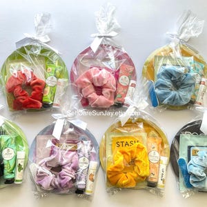 Puede incluir: Cestas de regalo con varios artículos de cuidado personal, que incluyen coleteros, bálsamo labial y lociones. Cada cesta está envuelta en celofán transparente y atada con una cinta blanca. Las cestas contienen artículos en tonos rojos, rosas, azules, verdes y amarillos.