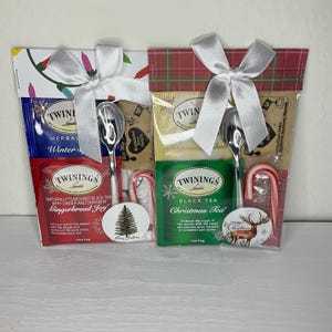 Puede incluir: Dos sets de regalo navideños, cada uno con una bolsita de té, una cuchara plateada, un bastón de caramelo y una pegatina. Uno tiene una bolsita de té azul y roja, el otro una verde, ambos atados con una cinta blanca.