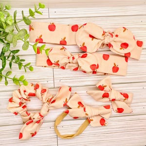 May include: A collection of peach-colored hair accessories with a red apple pattern. The set includes two headbands with bow accents, a scrunchie, and two bow clips. The accessories are arranged on a light wood-grain surface.