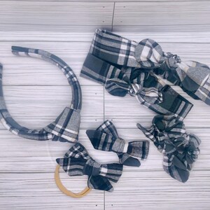 May include: A collection of baby accessories in a gray, black, and white plaid pattern. The set includes a headband, bow headbands, bow hair clips, and a scrunchie. The items are arranged on a light wood-grain surface.