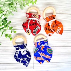 May include: Four baby teething rings with wooden handles and fabric designs. The fabric features college team logos in red, orange, blue, and navy. The logos include "Ohio State", "Penn State", and "Florida Gators".