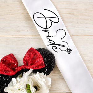 May include: A white satin sash with the word "Bride" in black script, featuring a Mickey Mouse head and a diamond ring graphic. Black sequined mouse ears with red sequined bows are visible, along with white flowers in a glass vase.