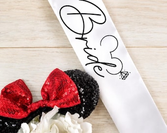 Puede incluir: Una banda de satén blanco con la palabra "Bride" en escritura negra, con una cabeza de Mickey Mouse y un gráfico de anillo de diamantes. Se ven orejas de ratón con lentejuelas negras con lazos de lentejuelas rojas, junto con flores blancas en un jarrón de cristal.