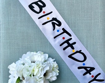 May include: White satin sash with the words "The One Where It's My Birthday" in black script with colorful dots.