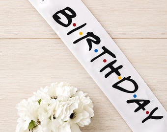 May include: White satin birthday sash with the text "The One Where It's My Birthday" and "BIRTHDAY" in black, with colorful dots. A bouquet of white flowers in a glass jar is in the lower left corner.