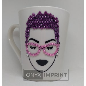 May include: White ceramic mug with a black and white illustration of a woman with purple and pink beaded hair and pink beaded glasses. The mug has the text "ONYX IMPRINT" printed on the bottom.