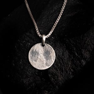 Pode incluir: Um pendente circular de tom prateado com uma superfície texturizada e irregular, suspenso por uma corrente de elos quadrados a condizer. O pendente apresenta uma pequena argola no topo, ligando-o à corrente metálica de padrão uniforme e constante.