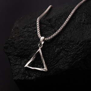 Puede incluir: Un collar con colgante plateado con un colgante triangular. El colgante tiene una superficie texturizada y cuelga de una cadena a juego. El collar se muestra sobre un fondo oscuro y texturizado.