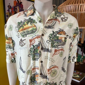 Pode incluir: Camisa creme com botões com estampa tropical com veleiros, palmeiras e as palavras "Changes in Attitudes" e "Margaritaville". A camisa de manga curta tem gola clássica e fecho de botões.