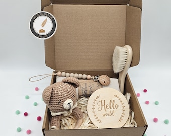 May include: A brown cardboard gift box containing a crocheted brown bear rattle, a wooden pacifier clip with light-colored beads, a wooden hairbrush with soft bristles, and a round wooden disc engraved with the text Hello world.