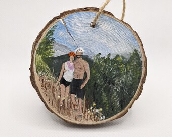 May include: A round wooden ornament with a painted scene of a couple hiking in a forest. The couple is wearing casual clothing and the woman is wearing a bright orange hat. The background is a blue sky with white clouds and green trees.