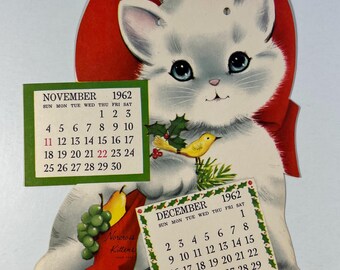 May include: A white kitten wearing a red bow holds a stocking with a pear and grapes. The kitten is holding a small yellow bird in its paw. The image includes a calendar for November and December of 1962.