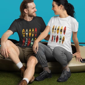 May include: A couple sit on a beige kayak with paddles. The man is wearing a black t-shirt with a colourful design of kayaks. The woman is wearing a white t-shirt with a colourful design of kayaks. They are both smiling and looking at each other.