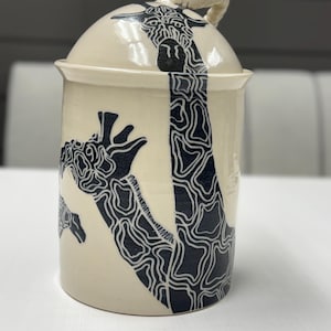 Könnte beinhalten: Cremefarbener Keramiktopf mit Deckel, mit einem schwarzen Giraffenmuster verziert. Das Giraffenmuster besteht aus einer Reihe schwarzer Linien. Der Deckel hat einen kleinen, gedrehten Seilgriff. Der Topf ist zylindrisch und steht auf einer weißen Oberfläche.