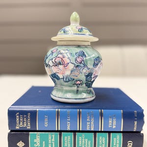May include: A small ceramic ginger jar with a lid, featuring a light green background and hand-painted pink and white floral motifs. The jar rests on two stacked blue Reader's Digest Select Editions books with gold lettering on the spines.