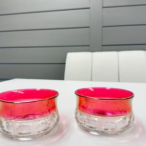 Könnte beinhalten: Zwei passende Glasschalen mit einem leuchtend pinkfarbenen oberen Bereich mit dünnem Goldrand und einem klaren, strukturierten unteren Sockel mit wiederkehrendem Prägemuster. Die Schalen haben eine weite Öffnung und eine runde, stabile Form.