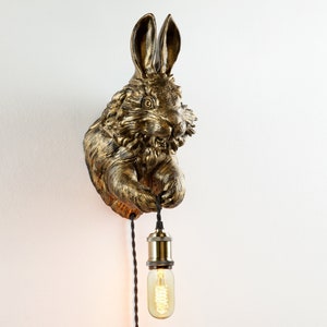 May include: A gold rabbit wall sconce with a hanging light bulb. The rabbit is depicted in a realistic style with fur and a long nose. The light bulb is a clear glass bulb with a filament.