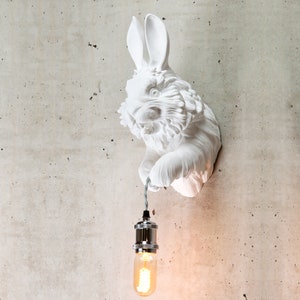 May include: White ceramic rabbit wall sculpture with a light bulb hanging from its paw. The rabbit has a textured fur and a realistic design.