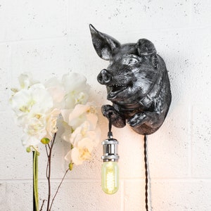 May include: A black and silver pig-shaped wall sconce with a light bulb hanging from its snout. The sconce is mounted on a white brick wall.