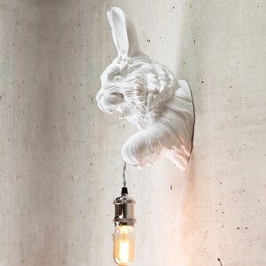 May include: White ceramic rabbit wall sconce with a hanging light bulb. The rabbit's head is turned to the side and its ears are perked up. The light bulb is a clear glass bulb with a silver metal socket.